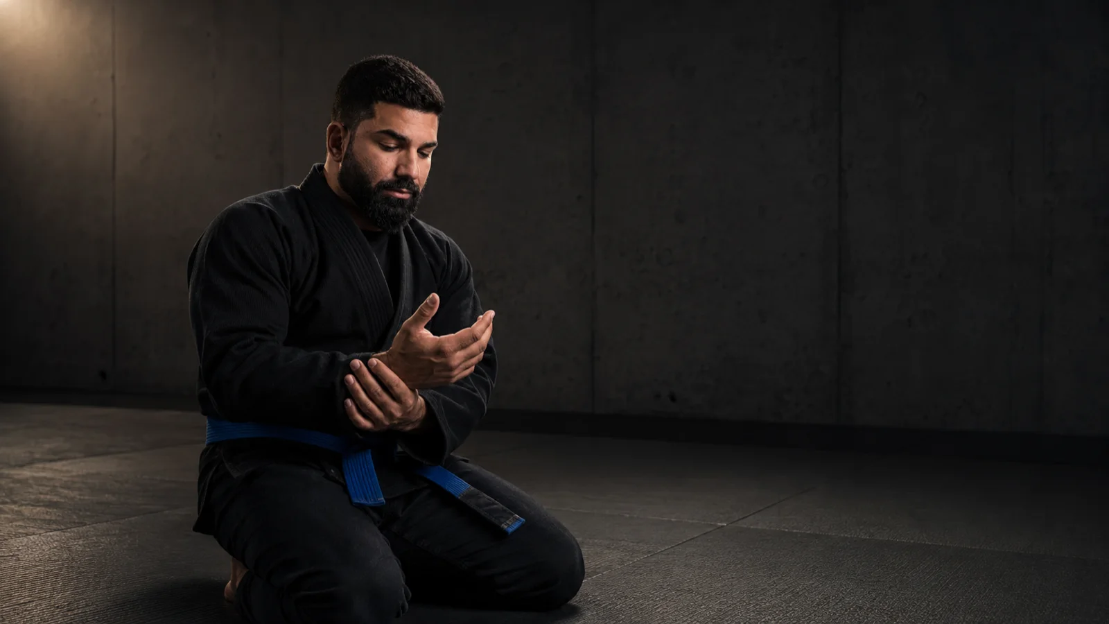 Pedro Dias ajoelhado no tatame de uma academia industrial, kimono preto e faixa azul, examinando a pegada antes do próximo rola